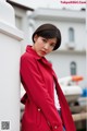 A woman in a red coat leaning against a white wall.