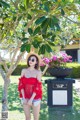 A woman in a red top and straw hat standing in front of a tree.